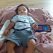 Cameron joined the competition — help win amazing prizes! baby, blanket, casual, child, clothing, curly_hair, cute, floor, indoor, lanyard, lying_down, person, playful, relaxed, shirt, shorts, toddler, toy_phone, wooden_floor, young