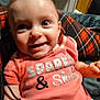 baby, child, smiling, pink_shirt, text_on_clothing, plaid_blanket, happy, indoor, face, person, cute, young_child, sitting, blue_eyes, short_hair, clothing, infant, portrait, casual, closeup