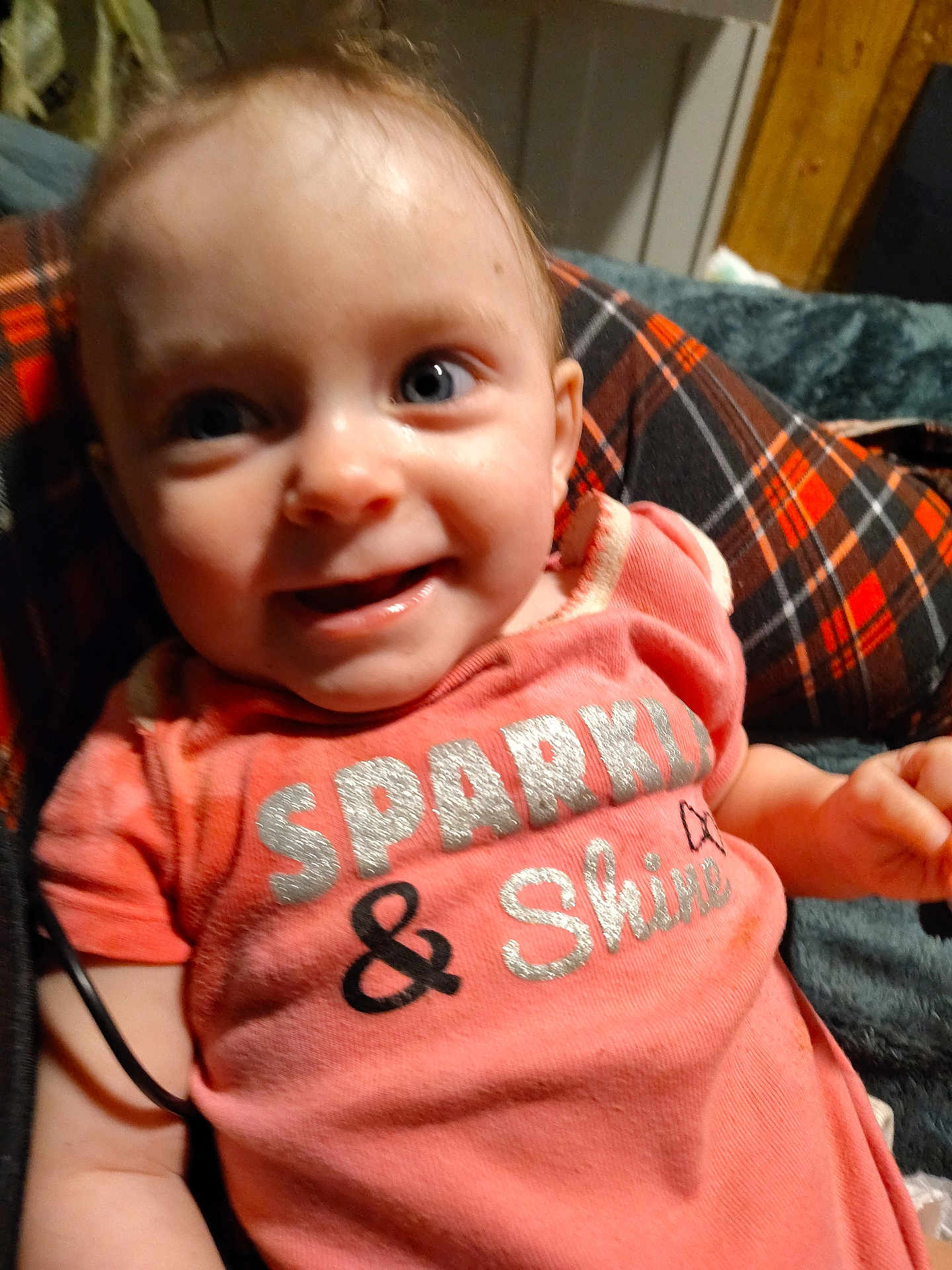 Maya is registered to the contest to win money with this photo: baby, child, smiling, pink_shirt, text_on_clothing, plaid_blanket, happy, indoor, face, person, cute, young_child, sitting, blue_eyes, short_hair, clothing, infant, portrait, casual, closeup