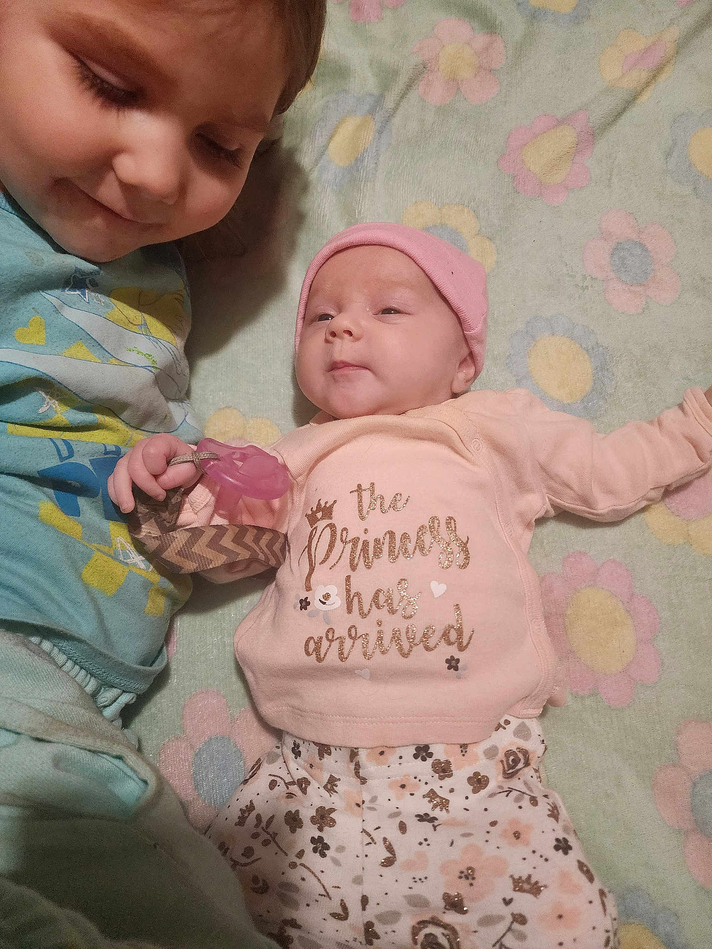 Harper is registered to the contest to win money with this photo: baby, child, newborn, sibling, smiling, cute, pacifier, pink_hat, floral_pants, blanket, lying_down, family, indoor, toddler, clothing, face, love, happy, portrait, infant