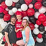 child, adult, family, graduation_cap, red_shirt, balloons, celebration, smile, glasses, tattoo, shorts, tshirt, indoor, floor, kneeling, posing, happy, people, party, background