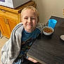 child, smiling, blanket, pajamas, table, bowl, pretzel_sticks, cup, kitchen, microwave, cabinet, wooden_cabinet, floor_tiles, chair, person, indoors, happy, young_child, cozy, snack