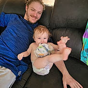 John is registered to the contest to win money with this photo: baby, man, couch, blue_shirt, diaper, blanket, smile, indoor, hardwood_floor, playful, relaxed, person, furniture, casual, home, child, kicking, happy, seated, light