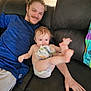 baby, man, couch, blue_shirt, diaper, blanket, smile, indoor, hardwood_floor, playful, relaxed, person, furniture, casual, home, child, kicking, happy, seated, light