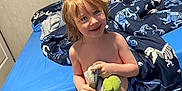 John joined the competition — help win amazing prizes! child, smiling, bedroom, bed, dinosaur_bedding, plush_toy, blanket, shorts, carpet, indoor, person, happy, hair, furniture, childhood, cute, casual, young, home, comfort