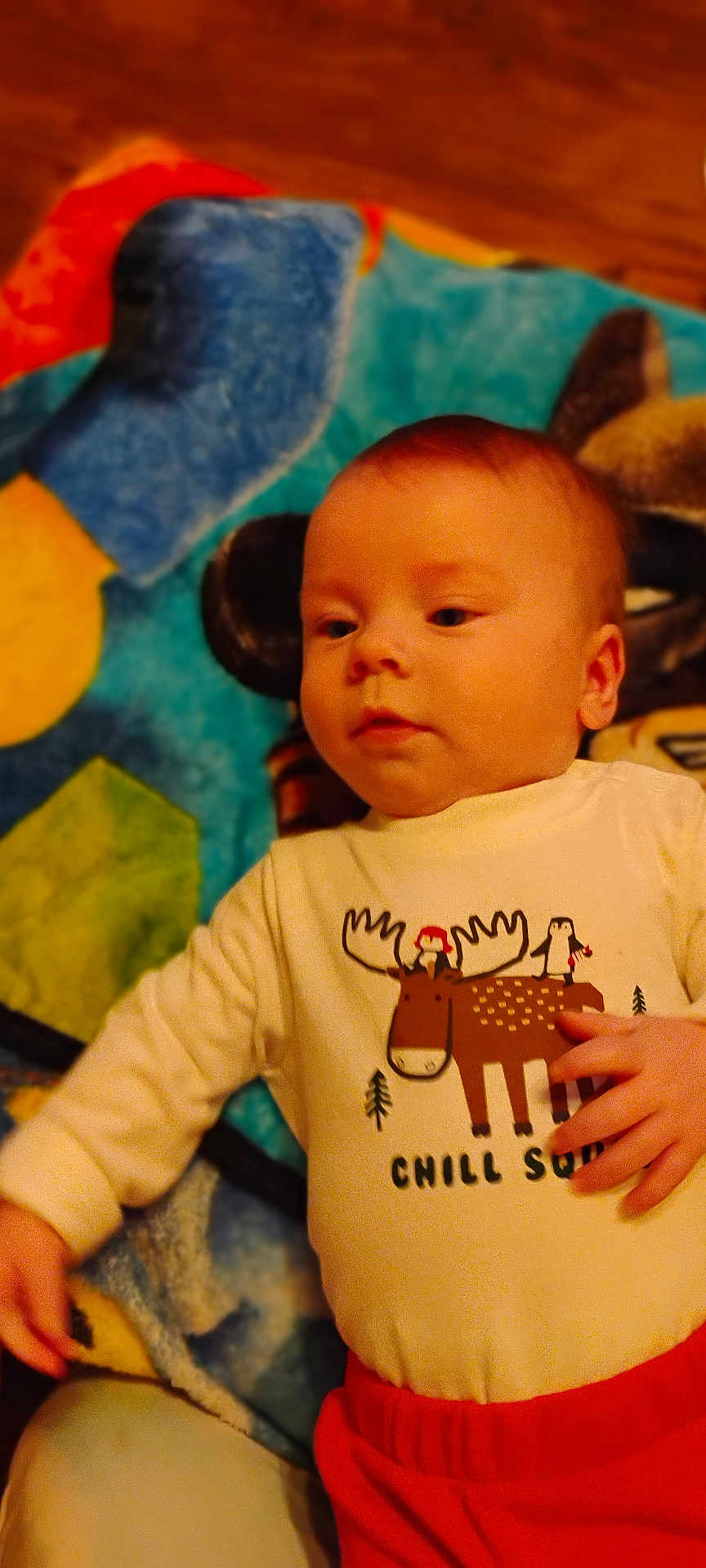 Asher is registered to the contest to win money with this photo: baby, child, infant, lying_down, shirt, graphic_tshirt, moose, penguin, blanket, colorful, red_pants, hand, face, head, cute, indoors, soft_texture, clothing, young, portrait