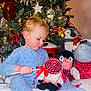 child, toddler, christmas_tree, plush_toy, holiday, pajamas, indoor, decorations, lights, ornaments, red_bow, stuffed_animal, festive, toy, winter, celebration, smiling, cute, cozy, family