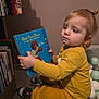 ball_pit, blond_hair, bookshelf, child, face, floor, hand, indoor, looking_right, picture_book, play_balls, portrait, reading, shoe, sitting, sneakers, soft_toys, toddler, wall, yellow_clothing