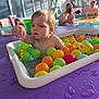 adult, baby, bath_toy, child, colorful_balls, curiosity, family, indoor_pool, parent, plastic_balls, plastic_container, playtime, poolside, purple_mat, splash, sunlight, swimwear, toddler, water, window