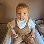 baby, baby_accessories, blond_hair, calm, child, cute, hands, indoor, infant, neutral_background, overalls, pacifier, person, portrait, sitting, skin, soft_surface, toddler, toes, white_bib