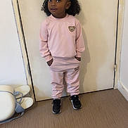 Inaya participe au concours pour gagner de l'argent avec cette photo : black_hair, casual_clothing, child, curly_hair, door, door_handle, floor, hands_in_pockets, indoor, looking_up, pants, pink_tracksuit, portrait, pose, sneakers, standing, sweatshirt, toddler, wall, young_child