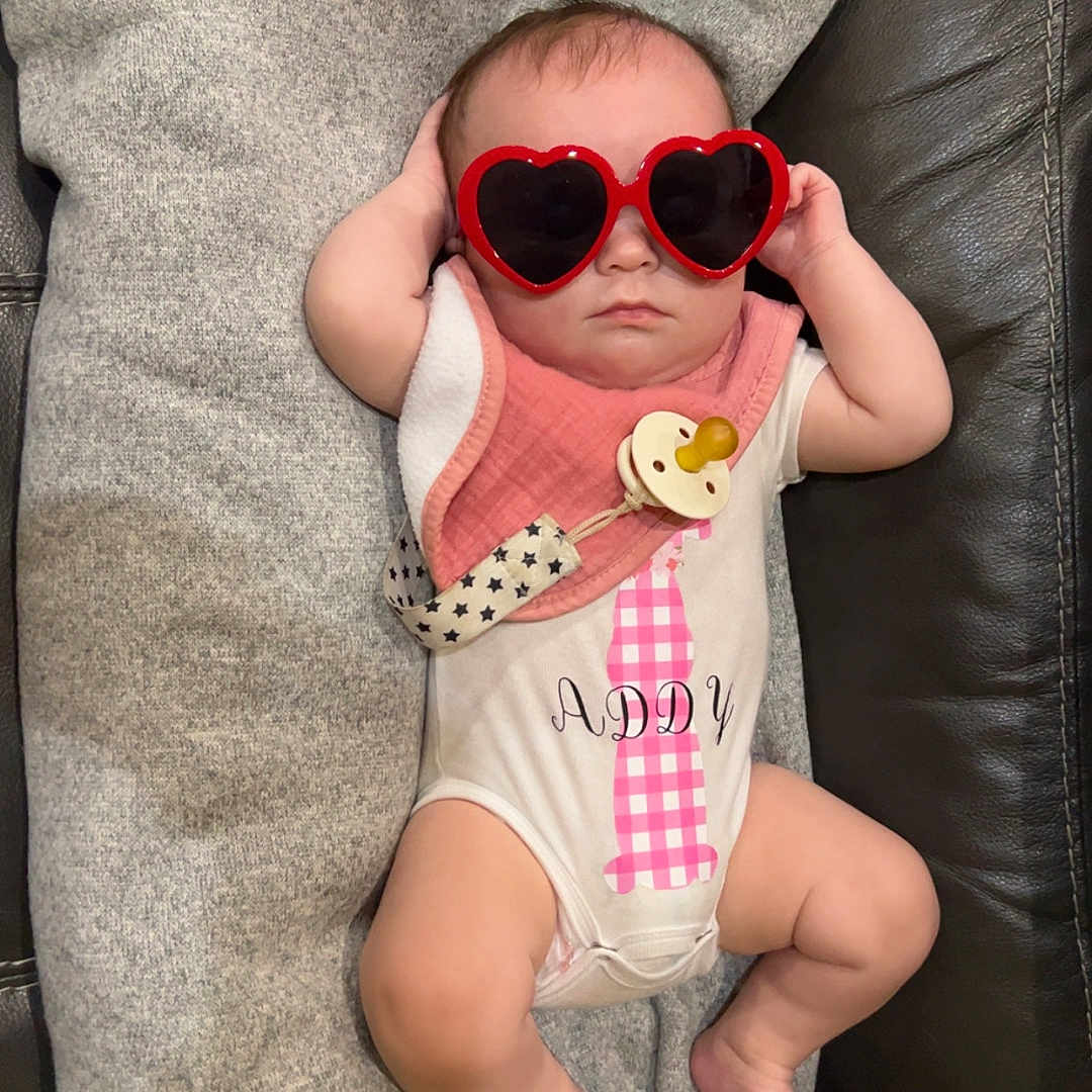 Addison joined the competition — help win amazing prizes! accessory, adorable, baby, baby_clothing, child, cushion, cute, fashion, funny, gray_blanket, heart_shaped, indoor, infant, leather_couch, onesie, pacifier, pink_bib, portrait, relaxed, sunglasses