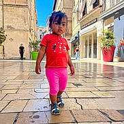 Athénaïs participe au concours pour gagner de l'argent avec cette photo : child, toddler, walking, street, pavement, buildings, outdoor, daylight, red_shirt, pink_leggings, sandals, people, urban, sidewalk, blue_sky, plants, shop, window, casual_clothing, curious