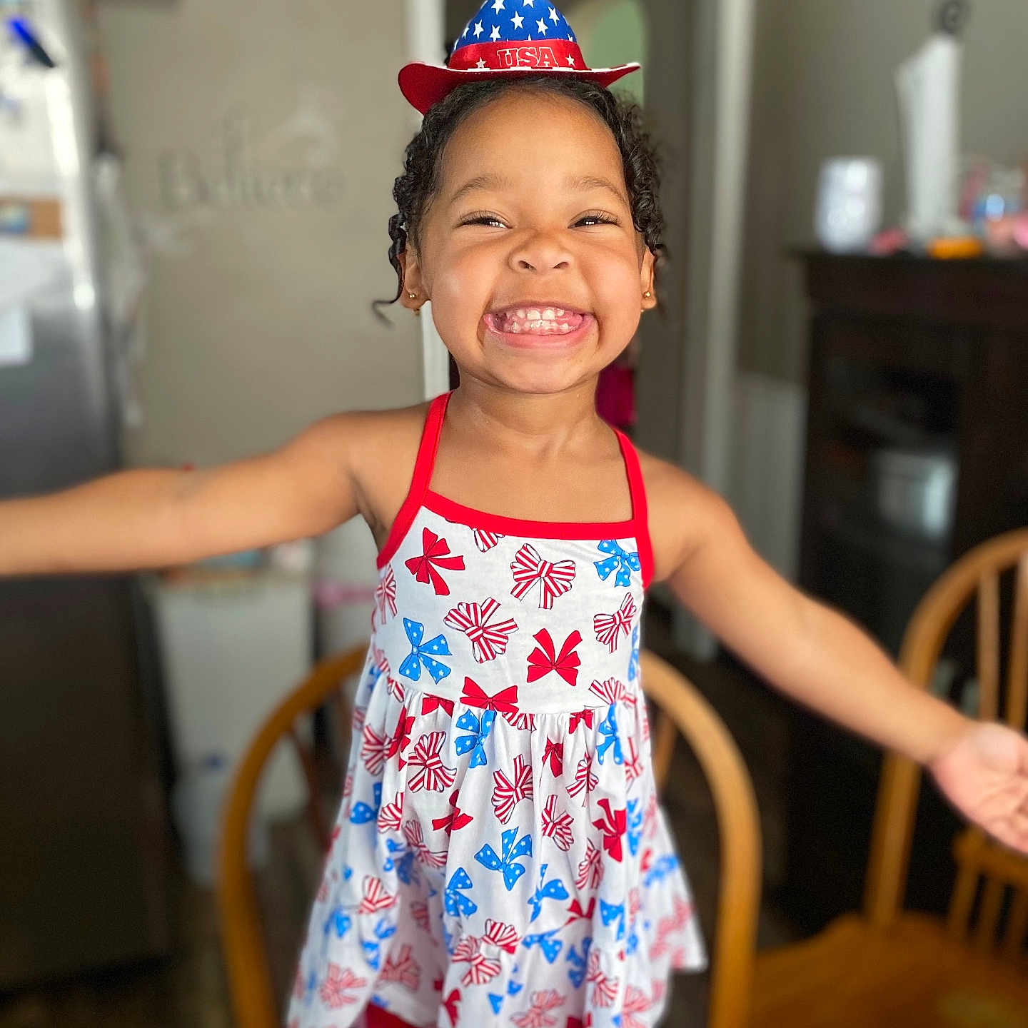 Sarai is registered to the contest to win money with this photo: blue, celebration, child, curly_hair, dress, furniture, girl, happy, hat, home, indoor, kitchen, patriotic, person, red, smile, standing, usa, white, wooden_chair