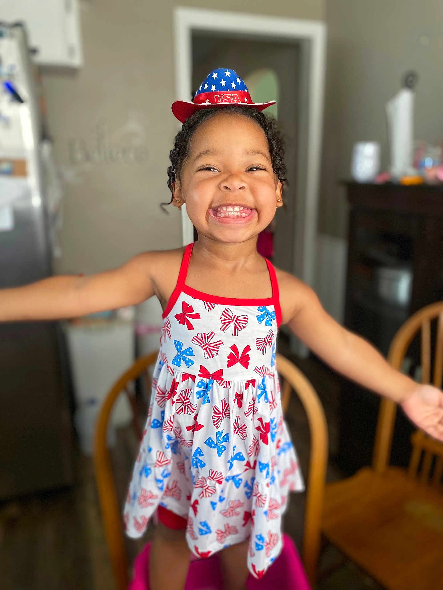 Sarai is registered to the contest to win money with this photo: child, girl, smile, dress, hat, usa, happy, indoor, kitchen, wooden_chair, patriotic, celebration, person, curly_hair, standing, red, white, blue, furniture, home