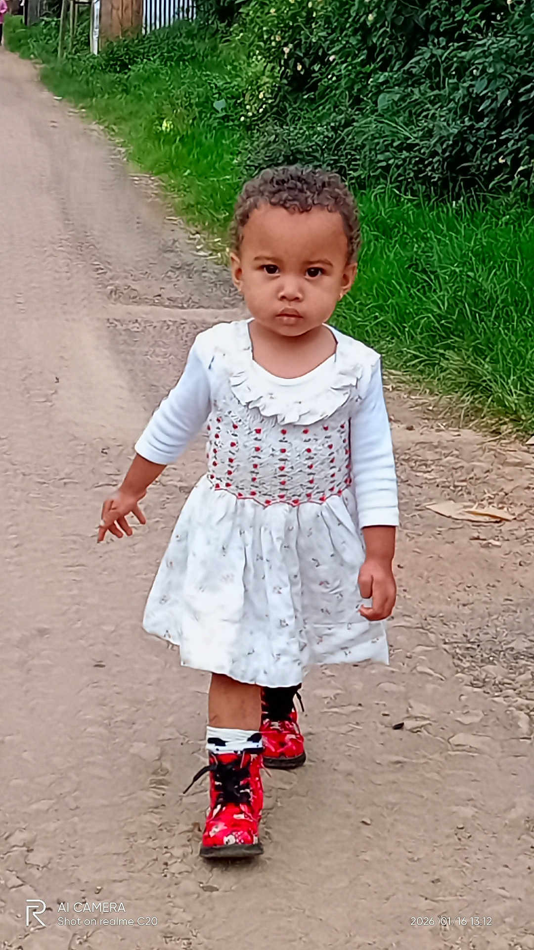 Almah a rejoint le concours — aidez-le/la à gagner de superbes lots ! casual_clothing, child, curly_hair, daytime, dirt_road, fence, footwear, grass, greenery, nature, outdoor, pathway, person, portrait, red_boots, serious_expression, toddler, walking, white_dress, young_child