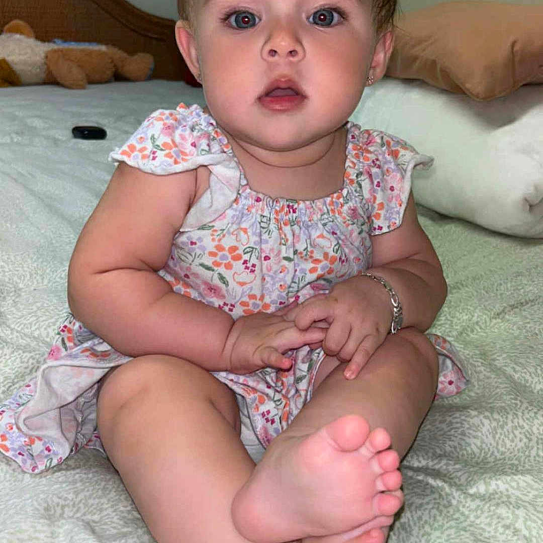 Blakeley is registered to the contest to win money with this photo: baby, barefoot, bed, bows, bracelet, child, curious, cute, face, floral_dress, hair_accessory, indoors, infant, person, pillow, portrait, sitting, skin, stuffed_animal, young