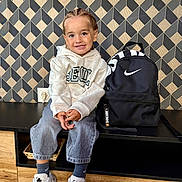 Swanny a rejoint le concours — aidez-le/la à gagner de superbes lots ! child, smiling, braided_hair, hoodie, jeans, sneakers, nike, backpack, black_backpack, modern_furniture, geometric_wall, indoor, floor_tiles, happy, seated, person, casual_clothing, portrait, cute, fashion