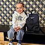 child, smiling, braided_hair, hoodie, jeans, sneakers, nike, backpack, black_backpack, modern_furniture, geometric_wall, indoor, floor_tiles, happy, seated, person, casual_clothing, portrait, cute, fashion