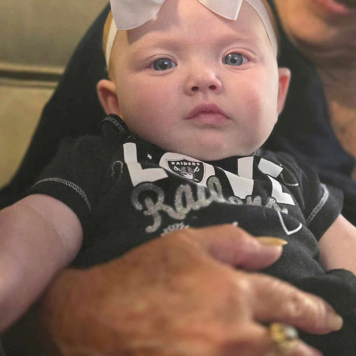 Deja Aznoe is registered to the contest to win money with this photo: baby, black_shirt, blurred_background, bow, candid, child, closeup, expression, face, hand, headband, holding, indoor, infant, older_adult, person, portrait, raiders, ring, smile