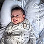 baby, smiling, face, onesie, paw_prints, tiger_patch, white_comforter, lying_down, infant, cute, happy, indoors, child, sleepwear, close_up, soft_lighting, portrait, small_hands, head, dark_hair