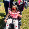 toddler, stroller, sunglasses, pacifier, pink_clothing, butterfly, heart_pattern, outdoor, grass, sunny, people, tent, shoes, child, casual, daytime, person, event, seat, fashion