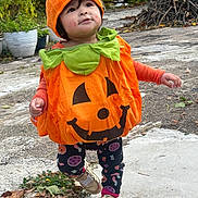 Jaylannie is registered to the contest to win money with this photo: child, toddler, pumpkin_costume, orange, green, outdoor, fall, leaves, sidewalk, plants, potted_plants, happy, cute, walking, shoes, leggings, playful, seasonal, costume, holiday