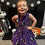 child, face_paint, purple_dress, halloween, ghost_decorations, skull_lights, plush_toy, carpet, smiling, pose, indoor, decorations, black_shoes, happy, festive, toy, costume, playful, home, celebration