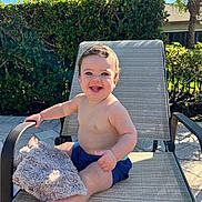 Jace is registered to the contest to win money with this photo: baby, child, smiling, outdoor, lounge_chair, blanket, happy, sunlight, greenery, palm_tree, shirtless, shorts, person, summer, vacation, relaxing, nature, feet, skin, portrait