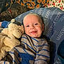 baby, smiling, stuffed_animal, blue_outfit, striped_clothing, cushion, dog, blanket, pumpkin_pattern, floral_pattern, indoor, cozy, child, person, happy, cute, portrait, lying_down, pet, soft_toys