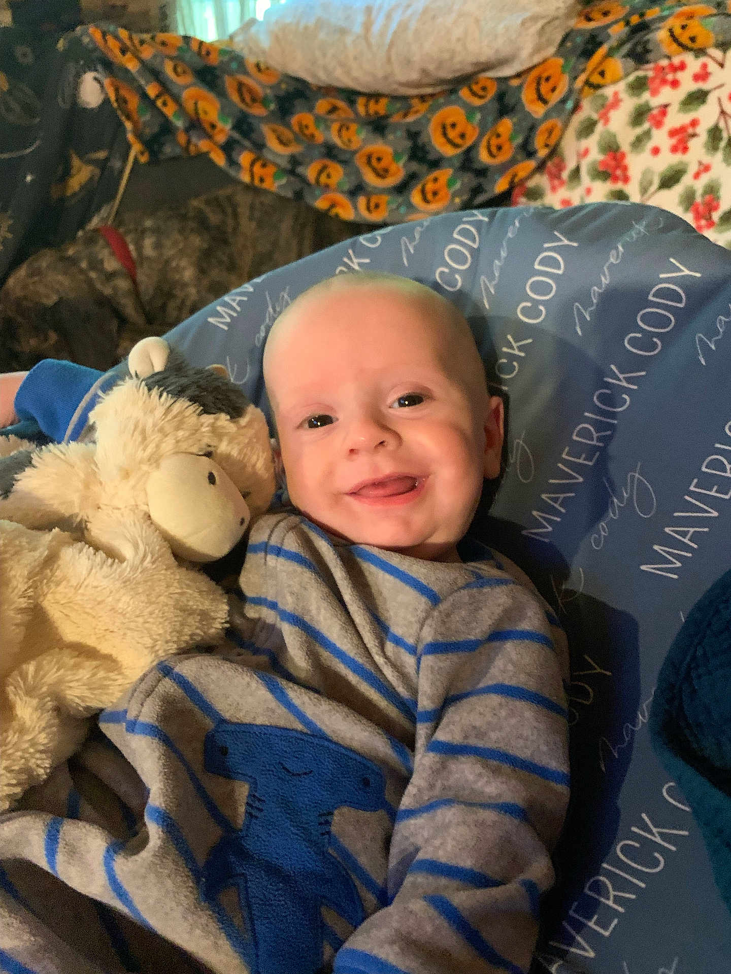 Maverick is registered to the contest to win money with this photo: baby, smiling, stuffed_animal, blue_outfit, striped_clothing, cushion, dog, blanket, pumpkin_pattern, floral_pattern, indoor, cozy, child, person, happy, cute, portrait, lying_down, pet, soft_toys
