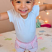 Aryia is registered to the contest to win money with this photo: toddler, child, smiling, pink_bow, curly_hair, white_shirt, pink_shorts, barefoot, bed, bedspread, animal_print, string_lights, indoor, happy, cute, portrait, playful, young_child, person, home