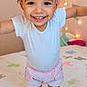 toddler, child, smiling, pink_bow, curly_hair, white_shirt, pink_shorts, barefoot, bed, bedspread, animal_print, string_lights, indoor, happy, cute, portrait, playful, young_child, person, home