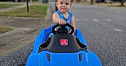 Colton is registered to the contest to win money with this photo: toddler, child, toy_car, blue_car, outdoor, street, residential_area, summer, daylight, trees, houses, sidewalk, playtime, cute, portrait, vehicle, road, happy, person, wheel