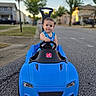 toddler, child, toy_car, blue_car, outdoor, street, residential_area, summer, daylight, trees, houses, sidewalk, playtime, cute, portrait, vehicle, road, happy, person, wheel