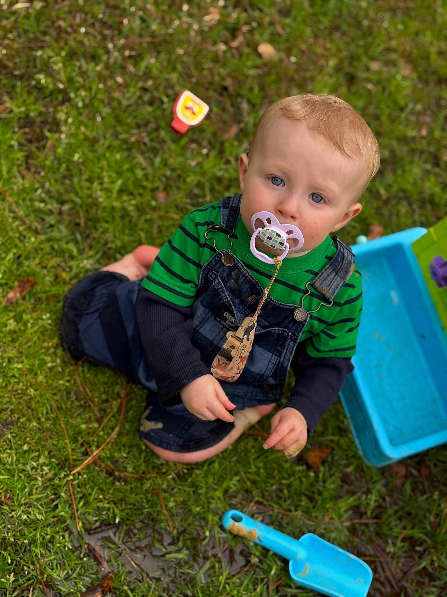 Ryder joined the competition — help win amazing prizes! child, baby, toddler, pacifier, overalls, striped_shirt, grass, shovel, sandbox, toy, blue_plastic, outdoor, sitting, mud, hands, face, blue_eyes, blond_hair, playtime, portrait