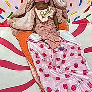 Amira is registered to the contest to win money with this photo: baby, child, pink_clothing, blanket, bib, hat, indoor, floor, decorations, colorful, arrows, strawberry_pattern, plush, cozy, portrait, seated, cute, young_child, warm, expression