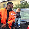 Kayden joined the competition — help win amazing prizes! man, child, toddler, smile, laughing, orange_jacket, nasa_patch, jeans, sneakers, car, license_plate, outdoor, daylight, person, holding, happy, urban, background, window, greenery