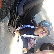 Kayden joined the competition — help win amazing prizes! child, boy, stroller, cap, toy, toy_gun, sidewalk, outdoor, smiling, casual_clothing, black_shoes, sunlight, playful, young, person, dinosaur_print, short_sleeve_shirt, black_and_white_shirt, concrete, happy