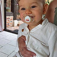 Santi participe au concours pour gagner de l'argent avec cette photo : toddler, child, pacifier, white_clothing, indoor, person, tattoo, hand, brick_wall, wood_beam, window, natural_light, floor_tiles, curtain, smile, face, baby, holding, sitting, cute