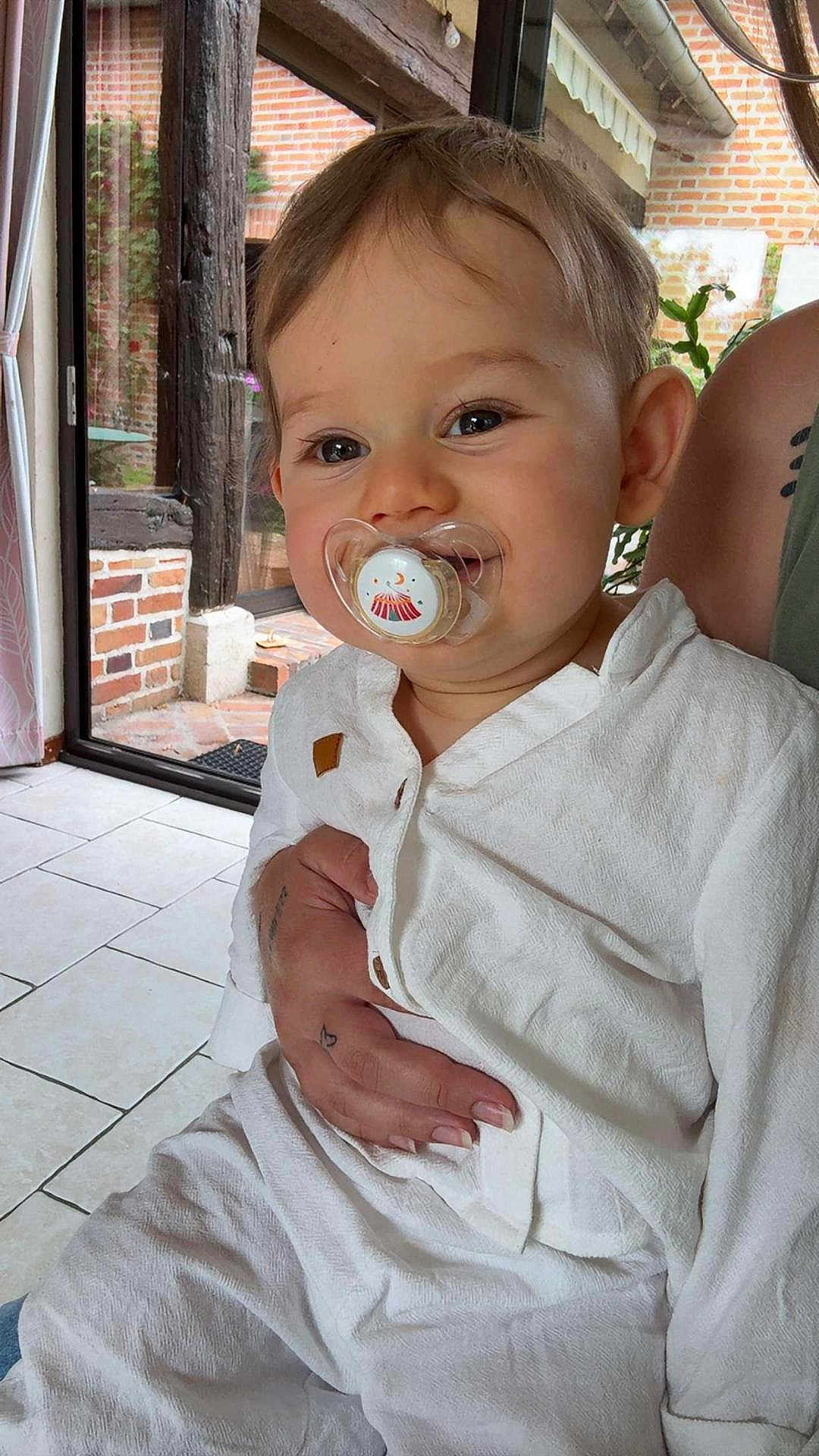 Santi participe au concours pour gagner de l'argent avec cette photo : toddler, child, pacifier, white_clothing, indoor, person, tattoo, hand, brick_wall, wood_beam, window, natural_light, floor_tiles, curtain, smile, face, baby, holding, sitting, cute