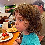 Mateo is registered to the contest to win money with this photo: child, indoor, restaurant, food, plate, table, person, brown_hair, turquoise_shirt, curly_hair, seated, beverage, adult, casual, dining, blurry_background, wooden_table, side_profile, focused, curious