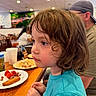 Mateo is registered to the contest to win money with this photo: adult, beverage, blurry_background, brown_hair, casual, child, curious, curly_hair, dining, focused, food, indoor, person, plate, restaurant, seated, side_profile, table, turquoise_shirt, wooden_table