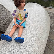 Mateo is registered to the contest to win money with this photo: child, toddler, portrait, sitting, park, pond, outdoor, sculpture, playground, trees, gazebo, water, blue_shoes, colorful_clothing, brown_hair, side_profile, long_legs, stone_pavement, daylight, recreation