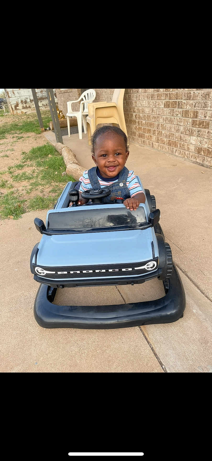 Jream joined the competition — help win amazing prizes! toddler, child, toy_car, blue, bronco, smiling, outdoor, concrete, driveway, brick_wall, plastic_chair, grass, fence, happy, person, steering_wheel, clothing, short_sleeves, overalls, daylight