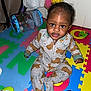 toddler, child, pajamas, bear_pattern, play_mat, toy, remote_control, colorful, indoor, floor, person, cute, curly_hair, sitting, baby, expression, plastic_toy, bag, home, playtime