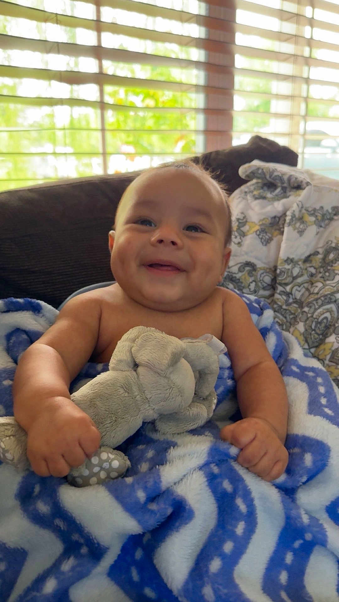 Waylon joined the competition — help win amazing prizes! baby, child, smiling, plush_toy, blanket, indoors, cozy, happy, cute, infant, furniture, couch, patterned_blanket, natural_light, window_blinds, portrait, skin, hand, face, expression