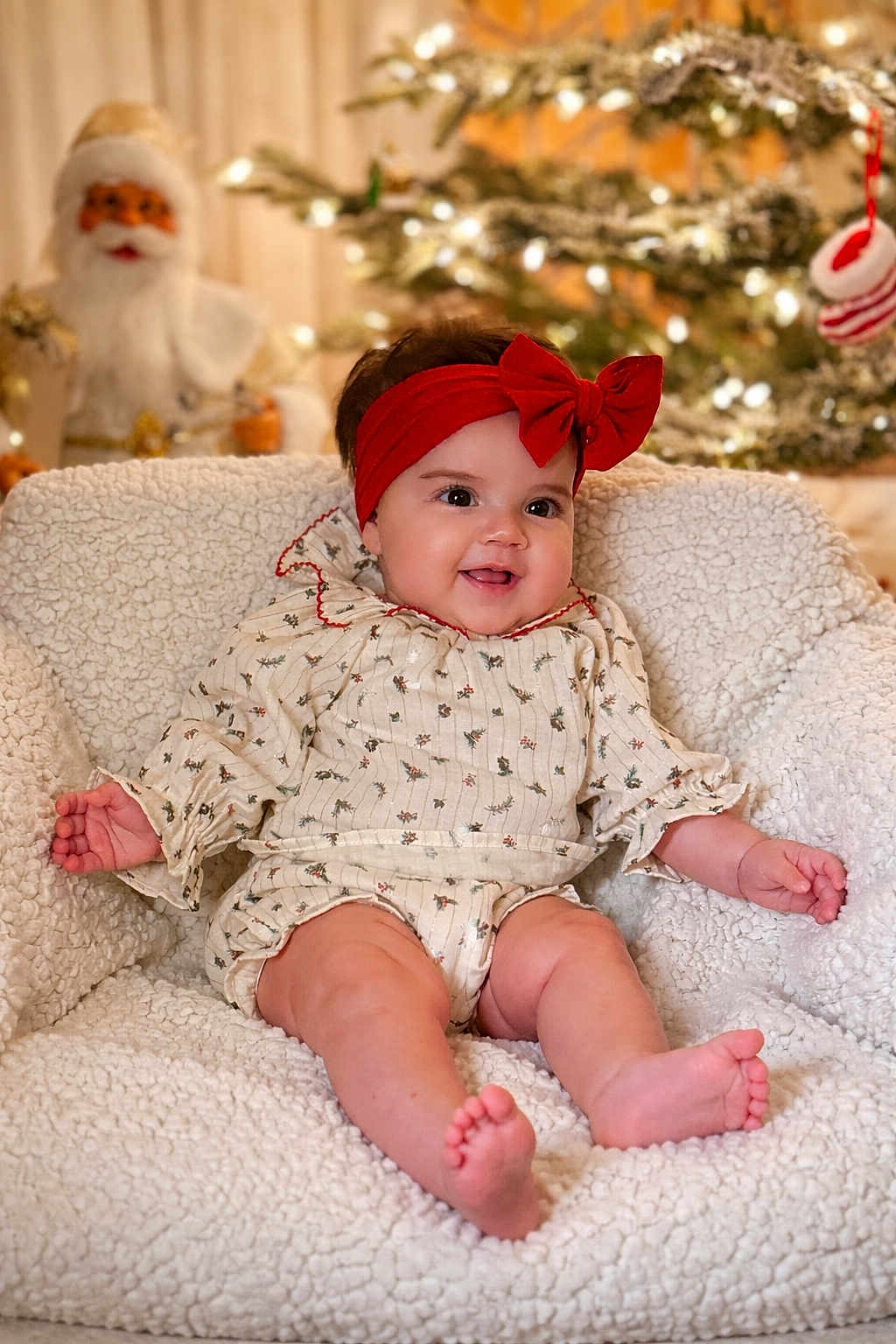 Amaia is registered to the contest to win money with this photo: baby, infant, red_headband, bow, white_chair, fluffy_texture, christmas_tree, holiday_decor, santa_decoration, smiling, cute, portrait, indoors, festive, seasonal, soft_lighting, child, happy, cozy, warm_colors