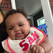 Jaliyah joined the competition — help win amazing prizes! baby, child, infant, bib, curly_hair, tongue_out, smiling, person, indoor, television, cartoon, hand, adult, brick_wall, doorframe, window, clothing, holding, face, cute
