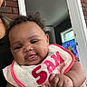baby, child, infant, bib, curly_hair, tongue_out, smiling, person, indoor, television, cartoon, hand, adult, brick_wall, doorframe, window, clothing, holding, face, cute